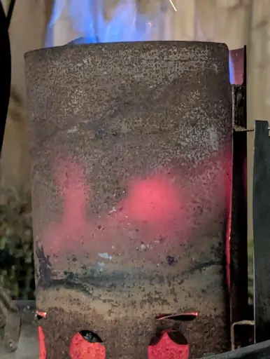 A close up photo of a rusty charcoal flume. The charcoal is lit, and has warmed up the sides of the flume to a dull red in patches. It is ready to be dumped into the grill and ready for food to start cooking over it.