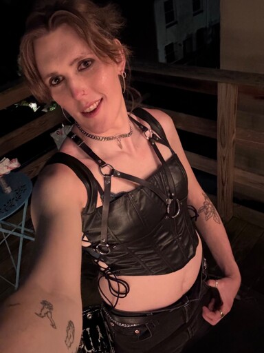 A selfie taken from a kind of high angle of a tall woman with dirty blond bangs, sparkly but alt makeup, a leather corset top that shows her midriff a bit with a leather harness over top and black cutoff jeans shorts. She’s wearing a chain choker and matching chain belt.