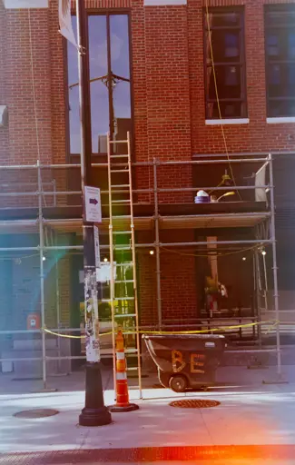 a film image of a bbrick building with scaffolding against it, and a ladder, and a bin. on the bin is taped the letters "BE". the image is affected by various streaks of light