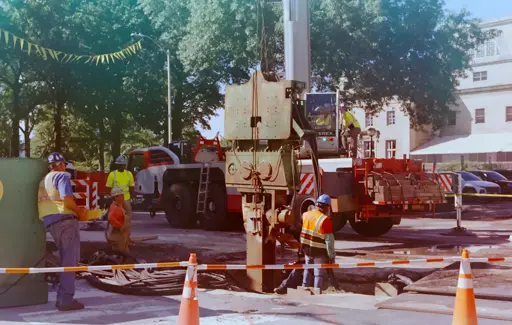 a film image of a group of constructions workers working around a large hole in the street. there is a large machine inserted into the street, of what type i dont know. 